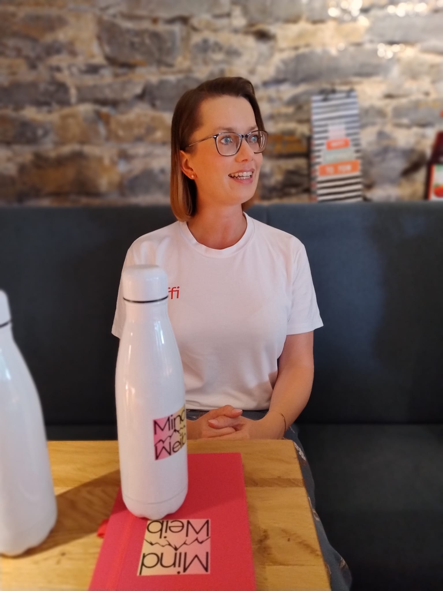Sophia Herrmann sitzt lächelnd an einem Holztisch beim MindWeib-Netzwerktreffen; im Vordergrund stehen eine MindWeib-Trinkflasche und ein pinkes Notizbuch.