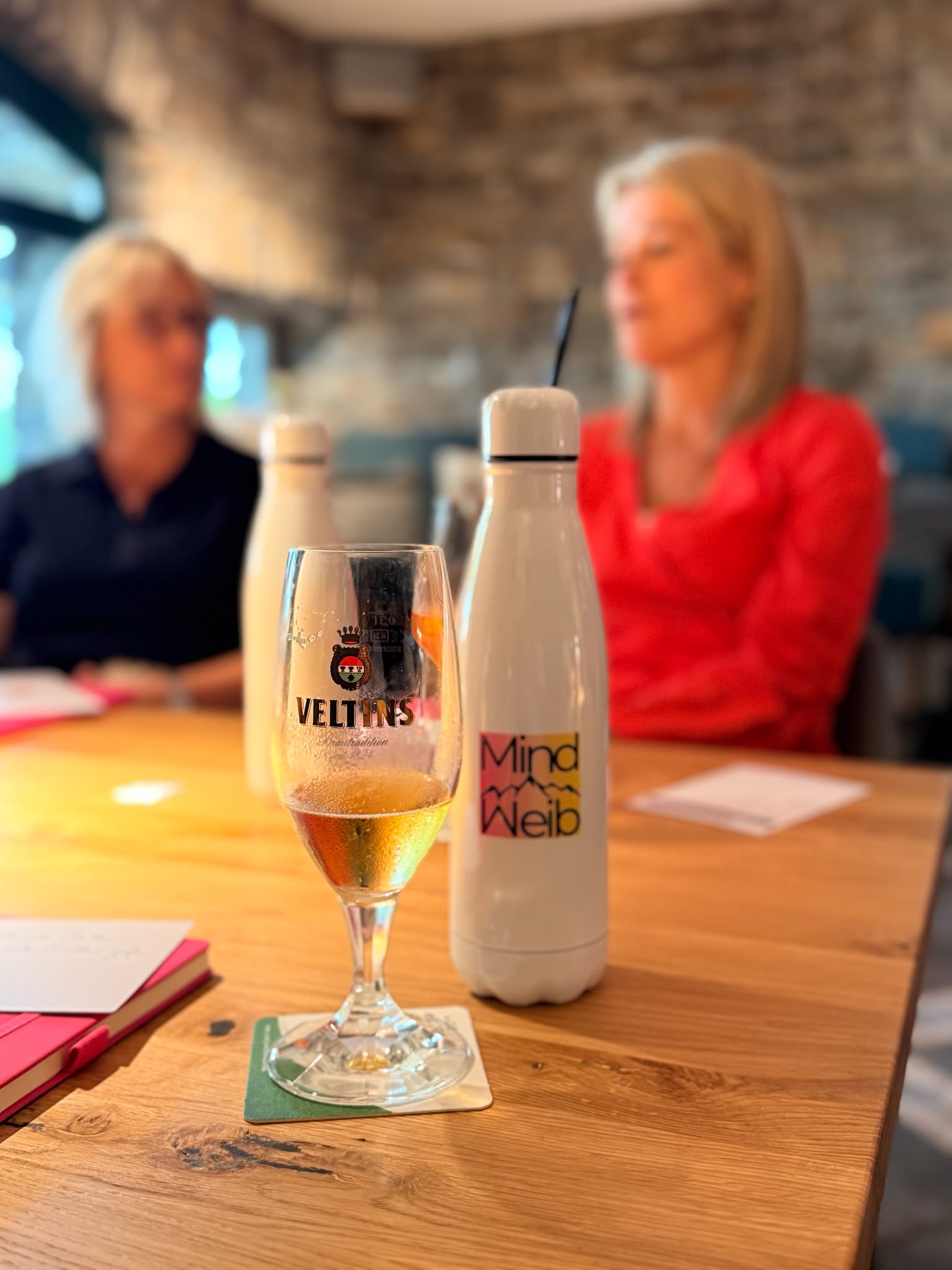 Veltins-Bierglas und weiße MindWeib-Trinkflasche auf einem Holztisch; im Hintergrund unterhalten sich zwei Frauen.