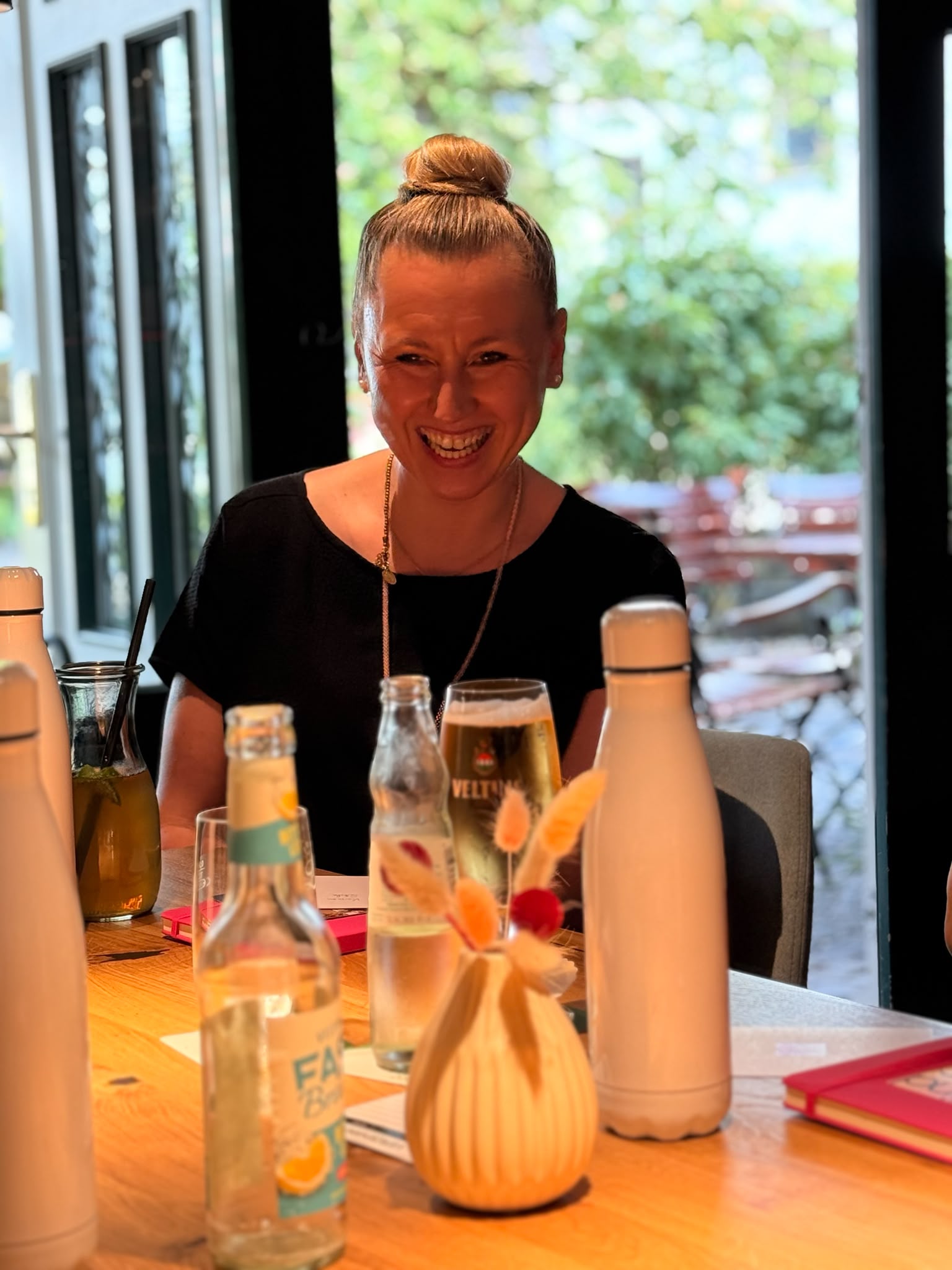 Lachende Frau am Holztisch beim MindWeib-Netzwerktreffen; vor ihr stehen Getränke, Trinkflaschen und Notizbücher.
