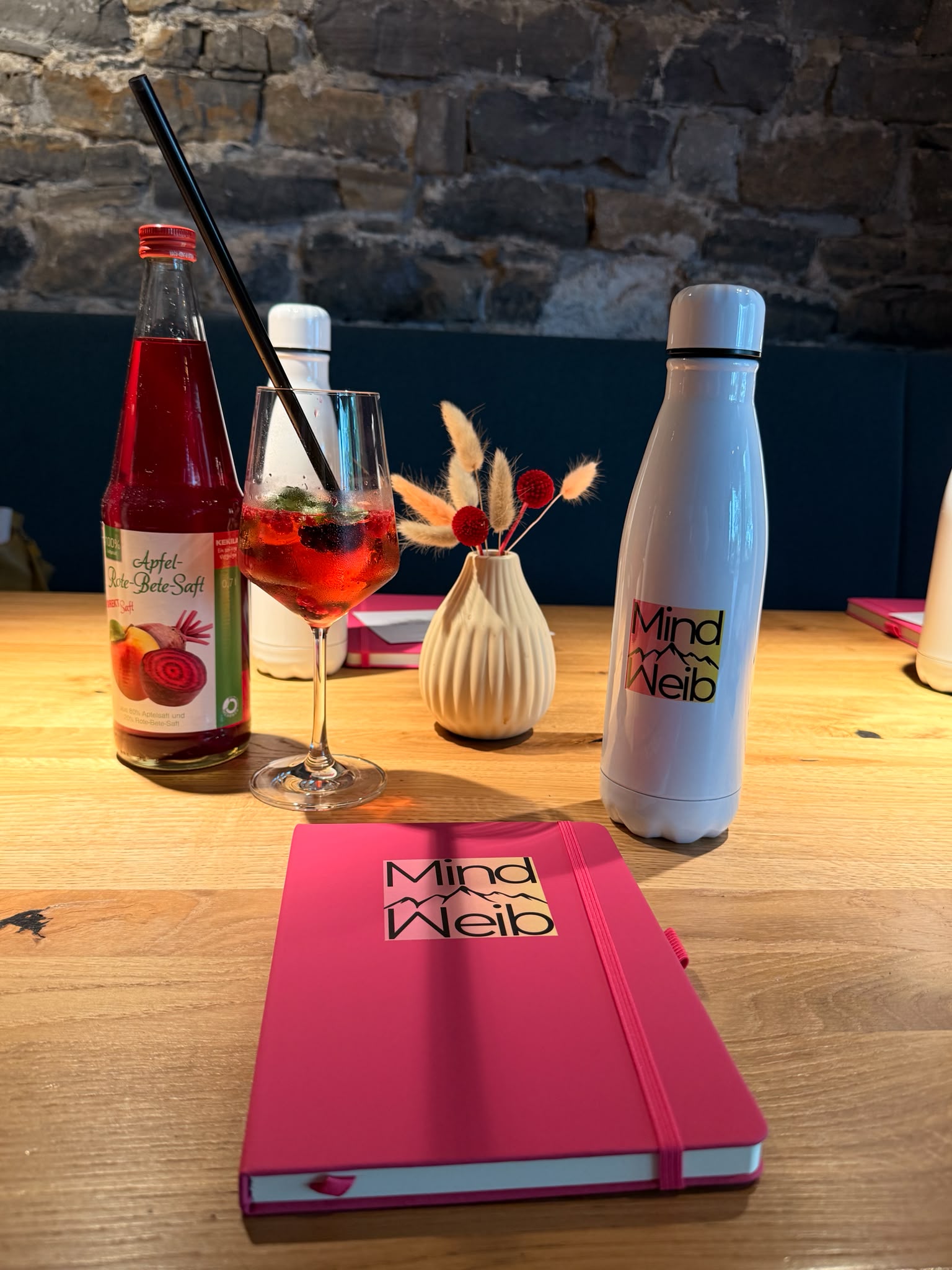 Pinks MindWeib-Notizbuch, weiße Trinkflasche, Getränk und kleine Vase auf einem Holztisch.