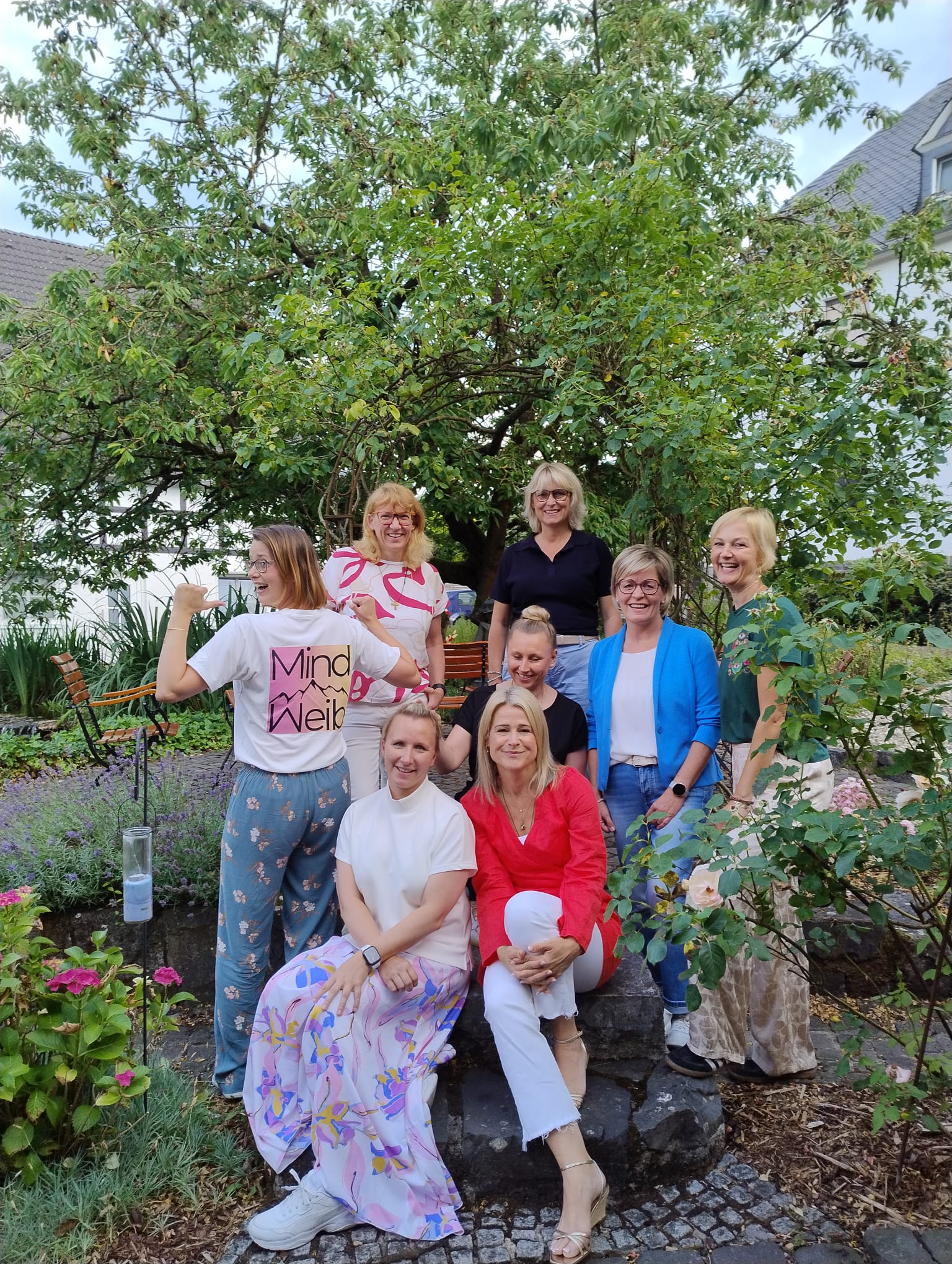 Acht Frauen aus dem MindWeib-Netzwerk posieren lachend in einem Garten; eine Frau im MindWeib-Shirt zeigt das Logo auf ihrem Rücken.
