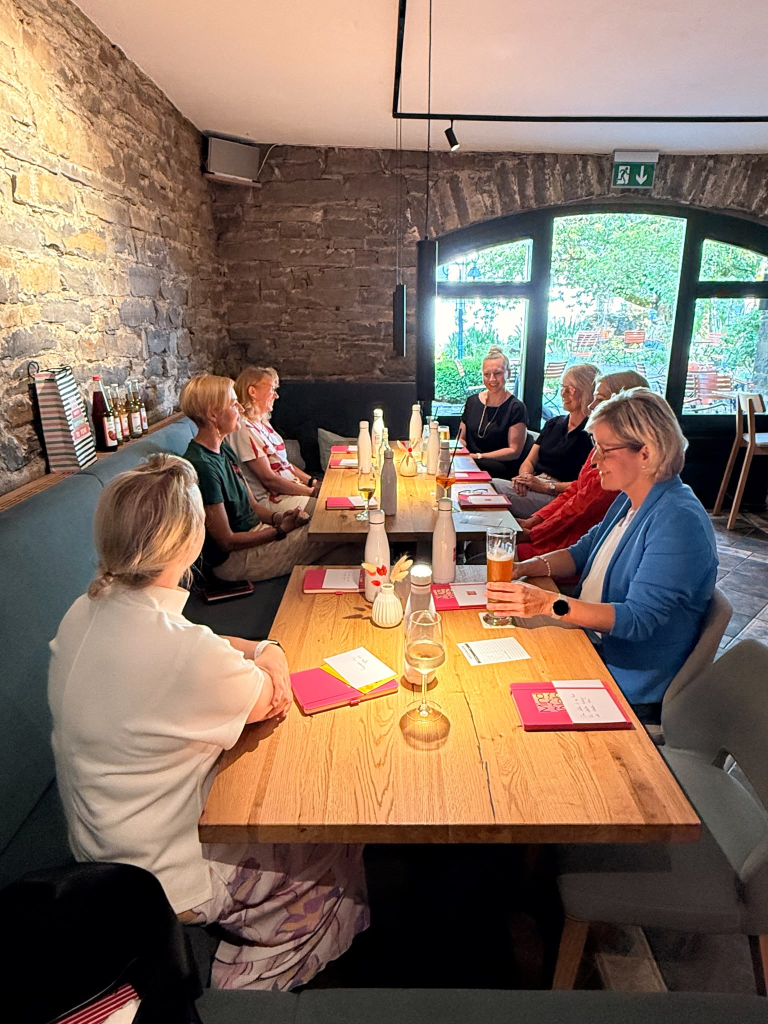 Sieben Frauen sitzen bei einem MindWeib-Netzwerktreffen an einer langen Holztischtafel in einem Restaurant mit Natursteinwand.