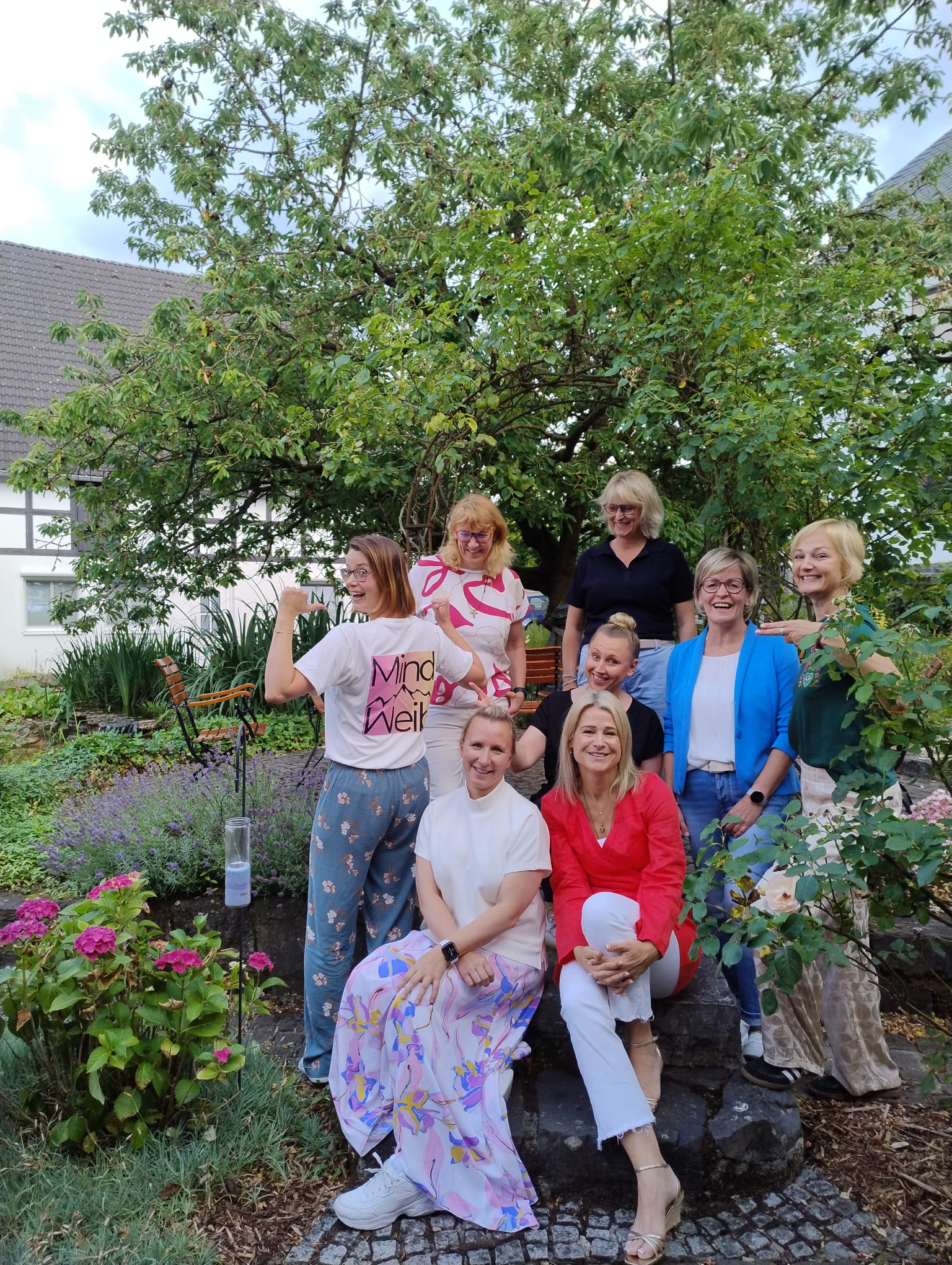 Acht Frauen aus dem MindWeib-Netzwerk stehen und sitzen für ein Gruppenfoto in einem blühenden Garten; eine Frau trägt ein MindWeib-Shirt.
